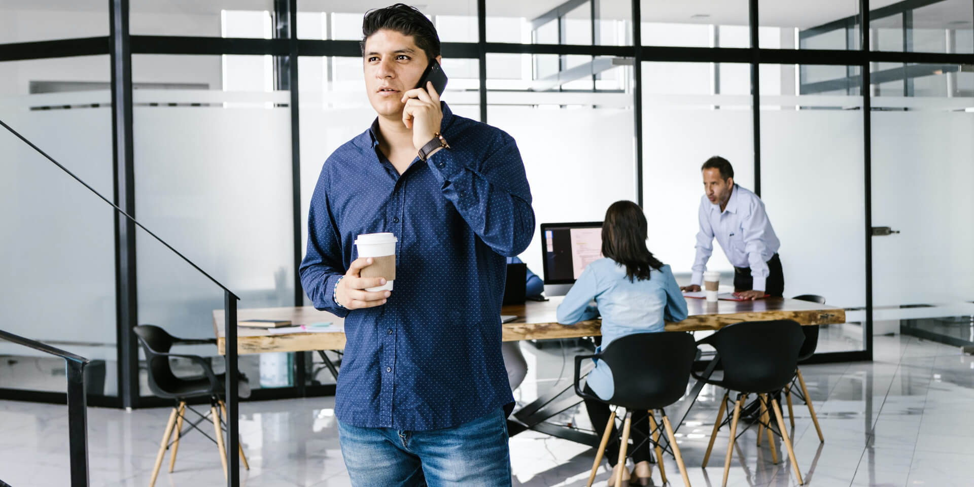 Een man in een blauw shirt staat in een modern kantoor, houdt een kopje koffie vast en praat op een mobiele telefoon. Op de achtergrond werken twee mensen aan een lange tafel met laptops. Het kantoor heeft grote glazen wanden en natuurlijk licht.