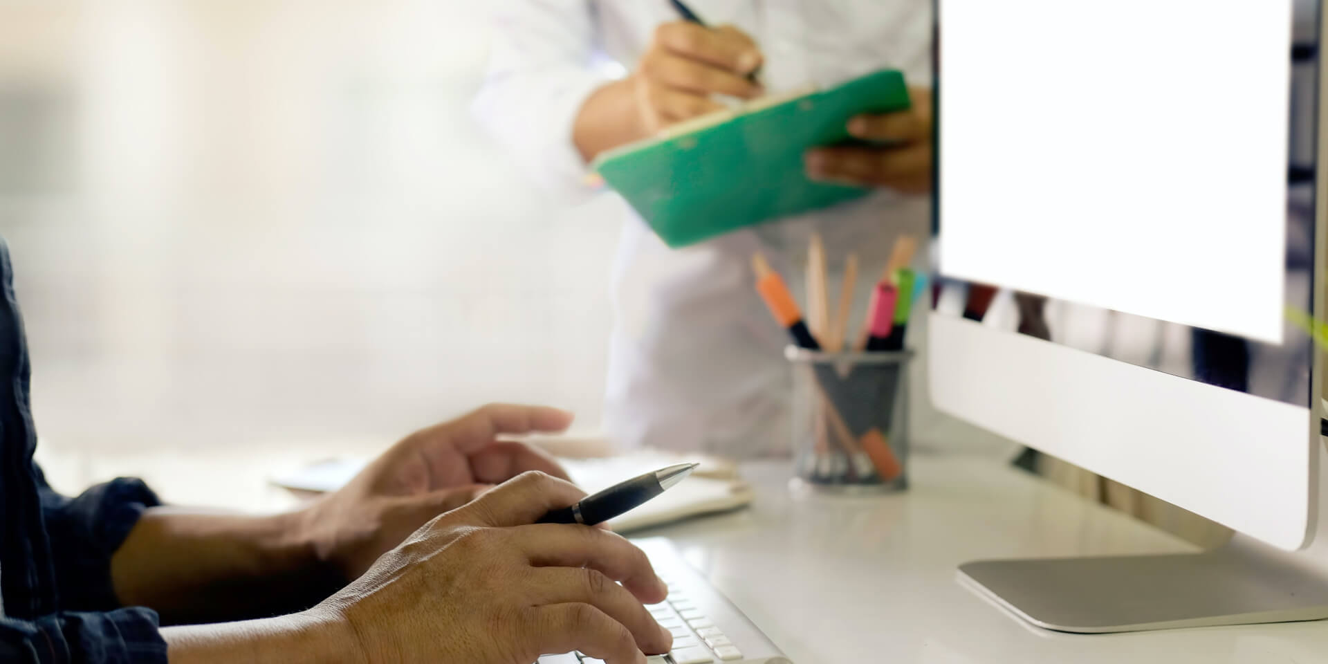 Twee mensen werken aan een bureau. De een gebruikt een computer met een tabletpen, terwijl de ander aantekeningen maakt op een klembord. Een beker bevat pennen en potloden naast de computer. De achtergrond is zacht vervaagd.