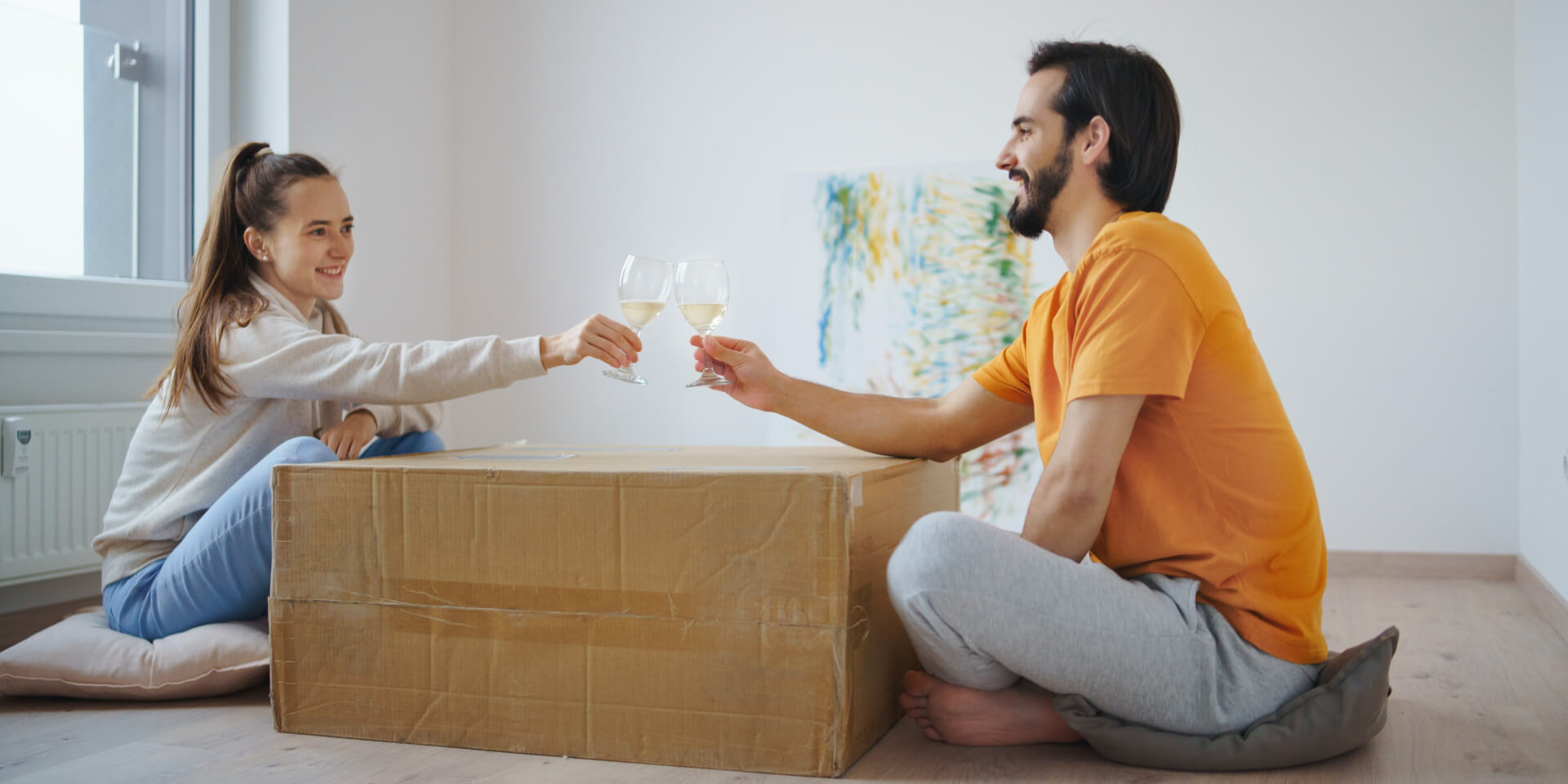 Een man en een vrouw zitten op kussens op de vloer en proosten met wijnglazen boven een grote kartonnen doos, die als geïmproviseerde tafel wordt gebruikt, in een felverlichte kamer. Een schilderij rust tegen de muur achter hen.