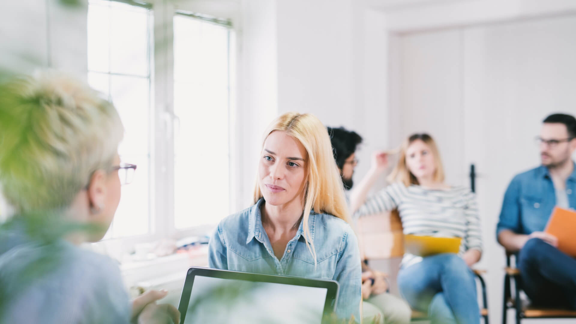 Een groep diverse mensen in een informele kantooromgeving. Een vrouw in focus praat met een persoon met kort haar. Anderen zitten en lijken betrokken, sommigen houden tablets en notitieboekjes vast. Zacht natuurlijk licht komt binnen via een raam.